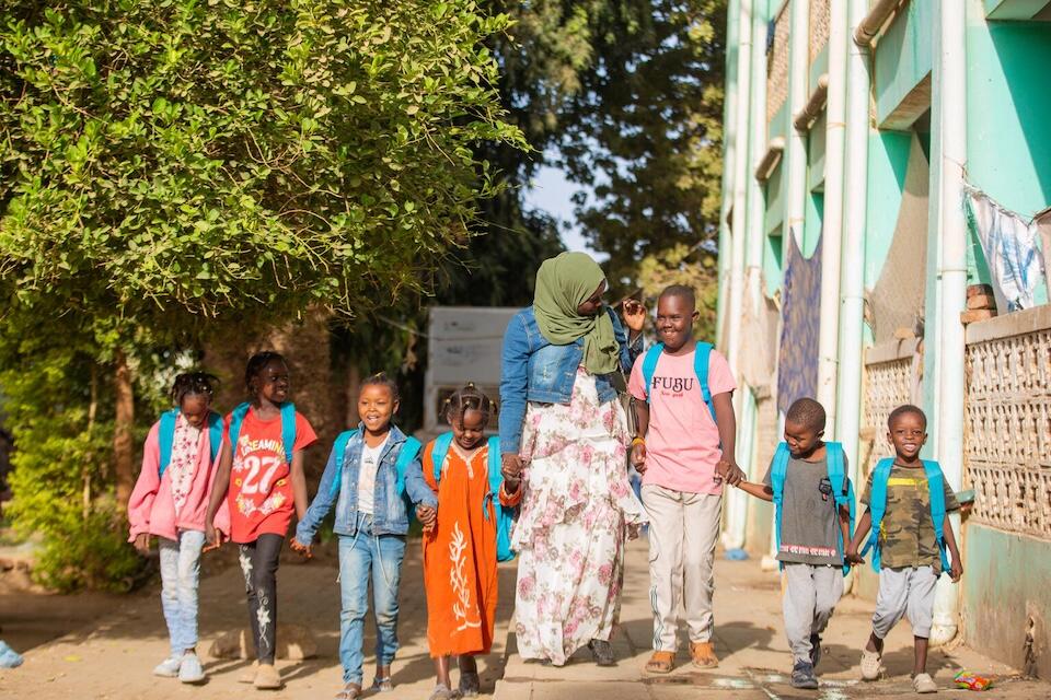Seven displaced children carrying UNICEF backpacks and their teacher head to the safe learning space UNICEF established at the At Al Salam displacement camp in Kassala state, Sudan.